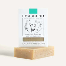 A white boxed Rosemary Mint Scrub milk soap with a goat illustration stands upright on a speckled tan soap bar.