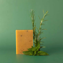 A rectangular golden-yellow bar of soap with small flecks stands upright beside fresh rosemary and mint sprigs against a green background.