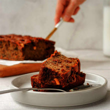 Two slices of dark banana bread rest on a white plate while a hand cuts the loaf behind.