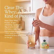 Woman holding glass of clear protein drink with Garden of Life Clear Whey Protein container on table beside her.
