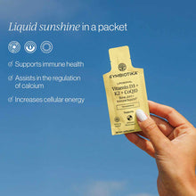 Hand holding a yellow Cymbiotika liposomal Vitamin D3+K2+CoQ10 supplement packet against a blue background.