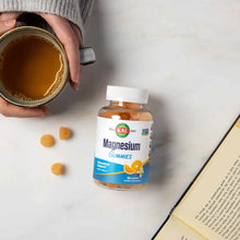Kal Magnesium Citrate Gummies bottle with white cap next to scattered orange gummies, cup of tea, and open book.