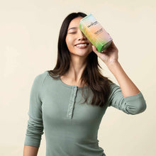 A smiling woman in a green henley top holds a colorful Hempress Organic Cover Panty Liners box against her forehead.