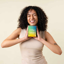 A smiling woman with curly dark hair holds a green and yellow box of Hempress Organic Cover Super Pads against a cream background.