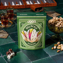 Joon dill pickle flavored pistachio package displayed on green tile with beverages, nuts in bowls, and gaming pieces.