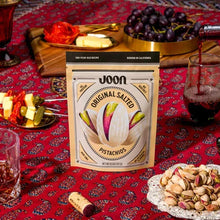 Joon brand original salted pistachios package on red patterned fabric surrounded by grapes, cheese, and nuts.