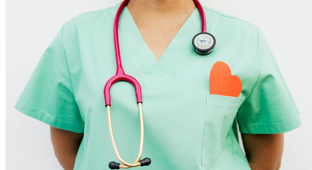 A healthcare worker in green scrubs with a stethoscope draped around their neck. A red paper heart is tucked into the chest pocket.