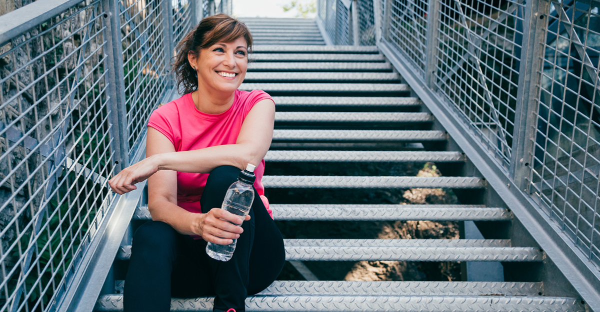 A woman in a pink shirt siting on metal stairs, smiling and holding a water bottle outdoors.