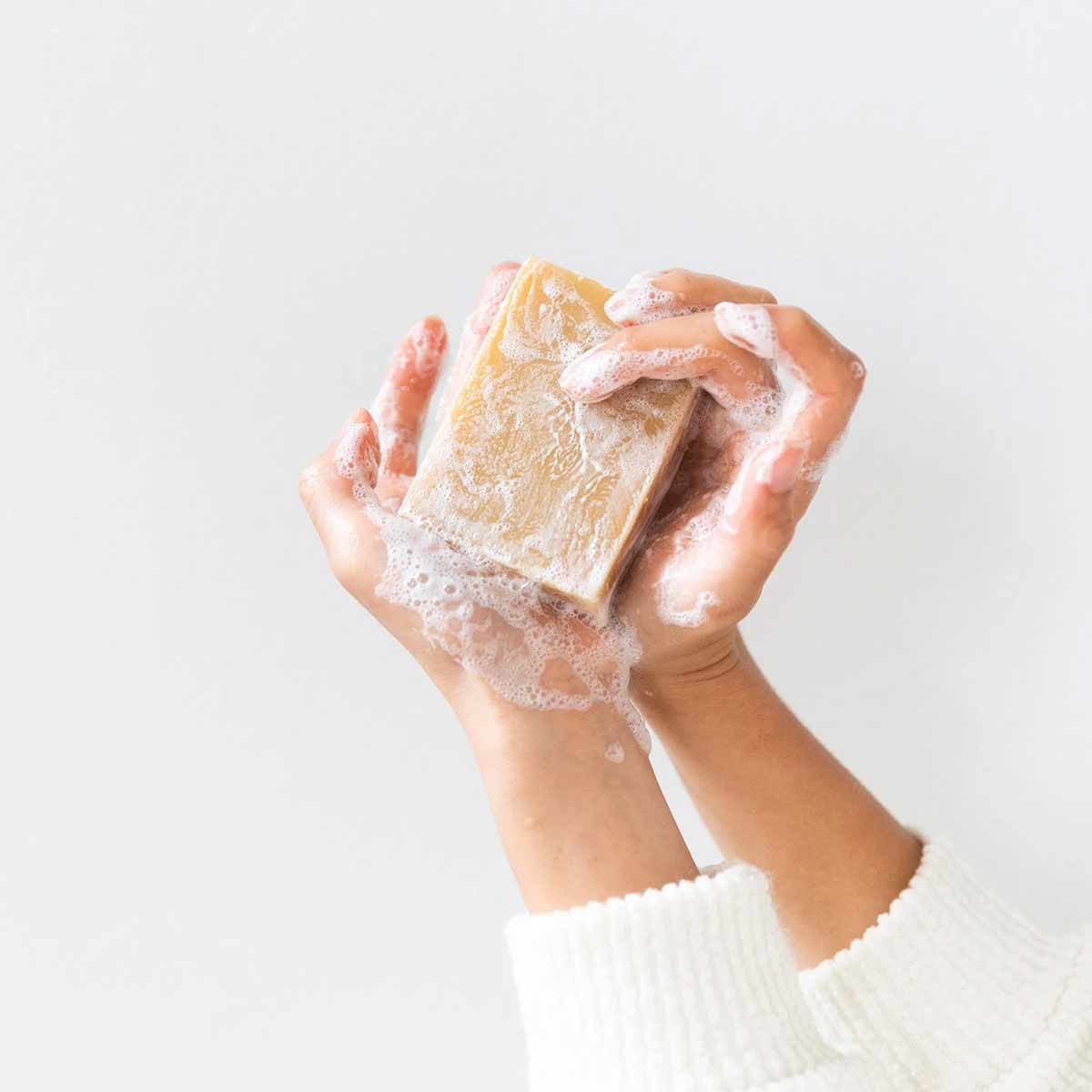 Two hands in a white knit sleeve hold a rectangular tan bar of soap covered in white lather against a plain background.