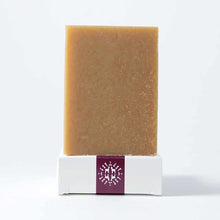 A rectangular tan bar of soap stands upright on a white box with a burgundy label against a plain background.