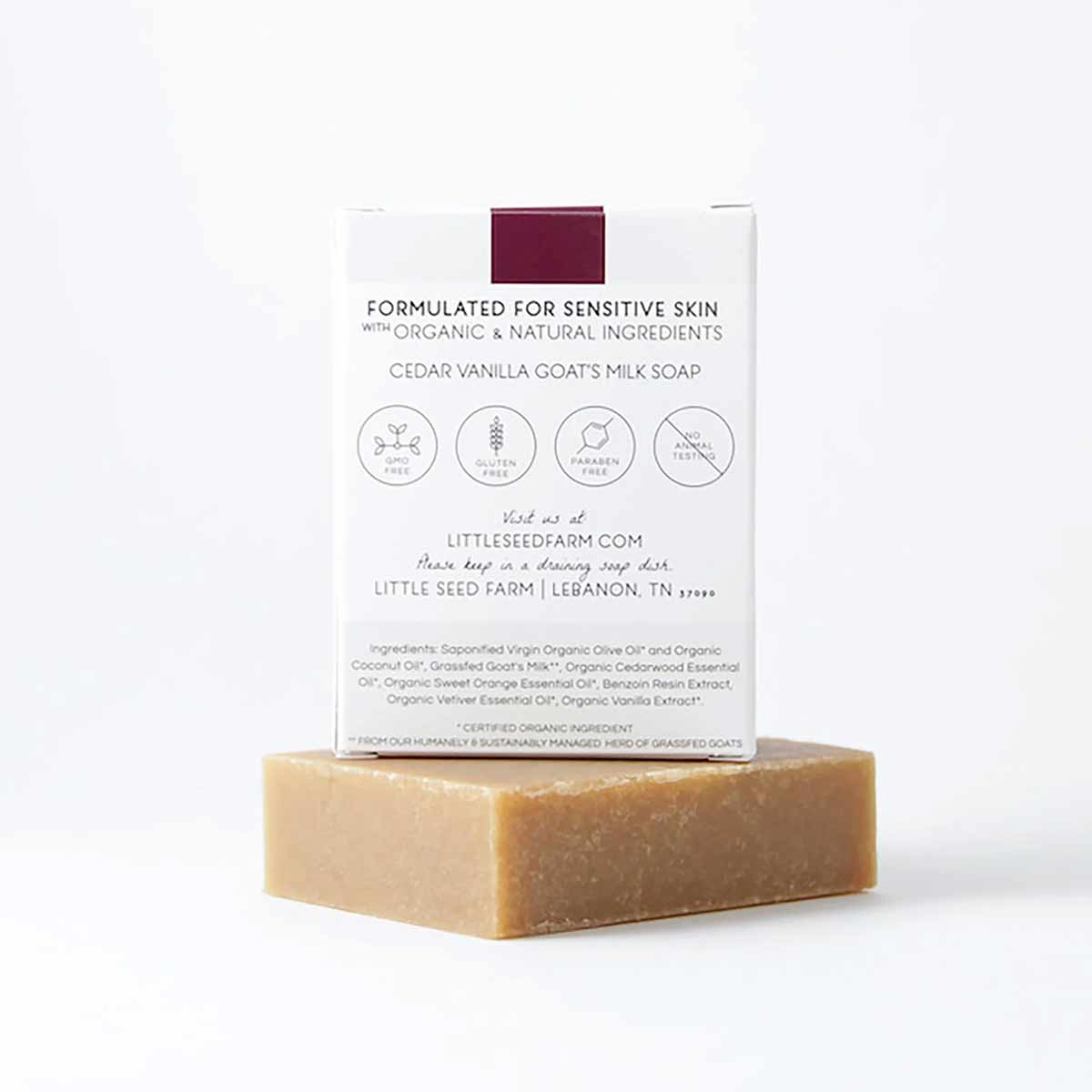 A tan bar of cedar vanilla goat's milk soap supports a white box with a maroon stripe and ingredient text.
