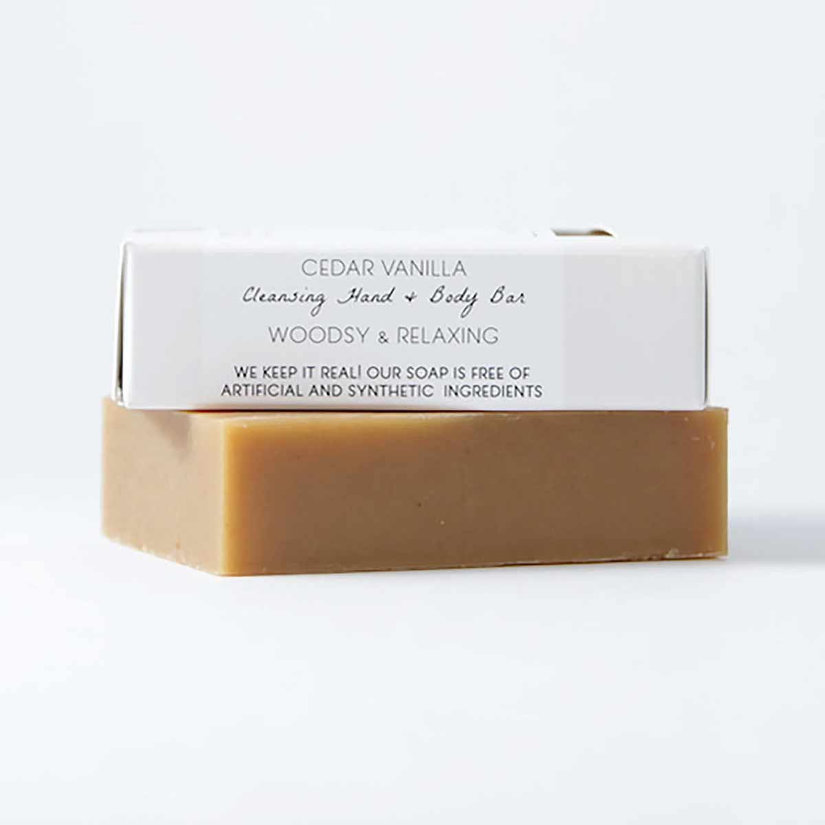 A tan rectangular soap bar sits beneath a white paper wrapper labeled Cedar Vanilla Cleansing Hand and Body Bar.