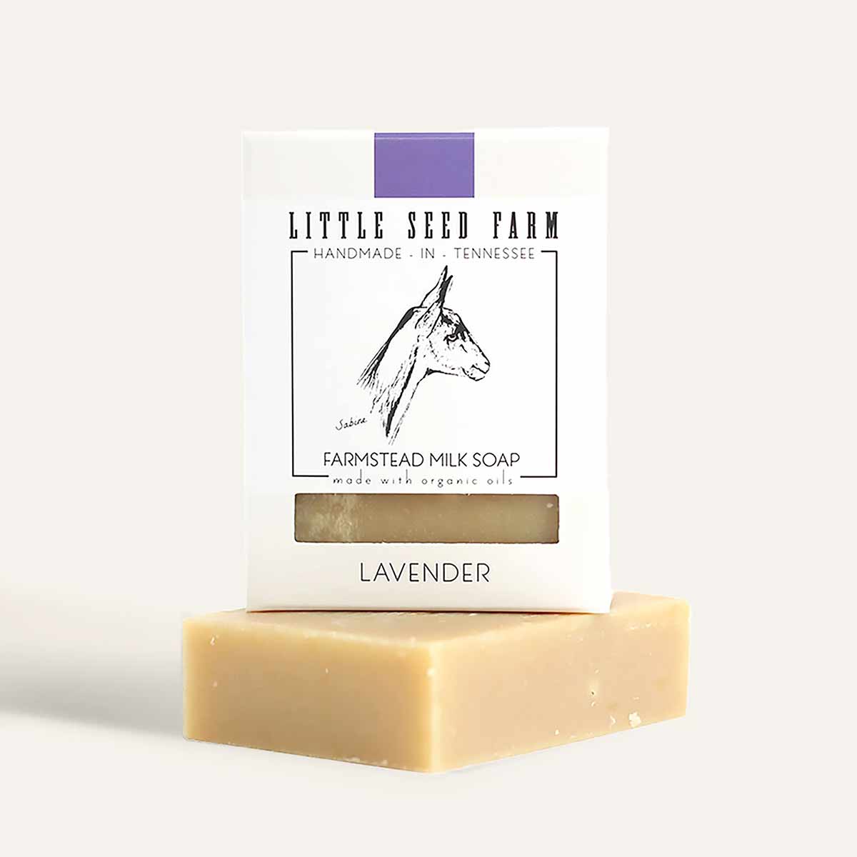 A cream-colored bar of lavender milk soap rests beneath a white Little Seed Farm package with a purple stripe and goat illustration.