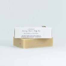A beige bar of soap sits beneath a white paper wrapper labeled Lavender Cleansing Facial and Body Bar against a plain background.