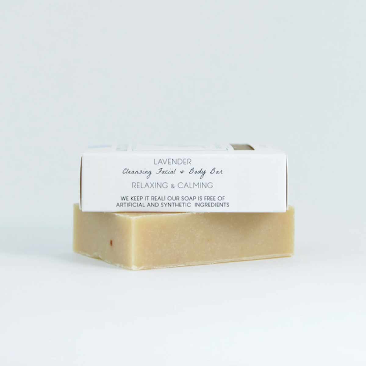 A beige bar of soap sits beneath a white paper wrapper labeled Lavender Cleansing Facial and Body Bar against a plain background.