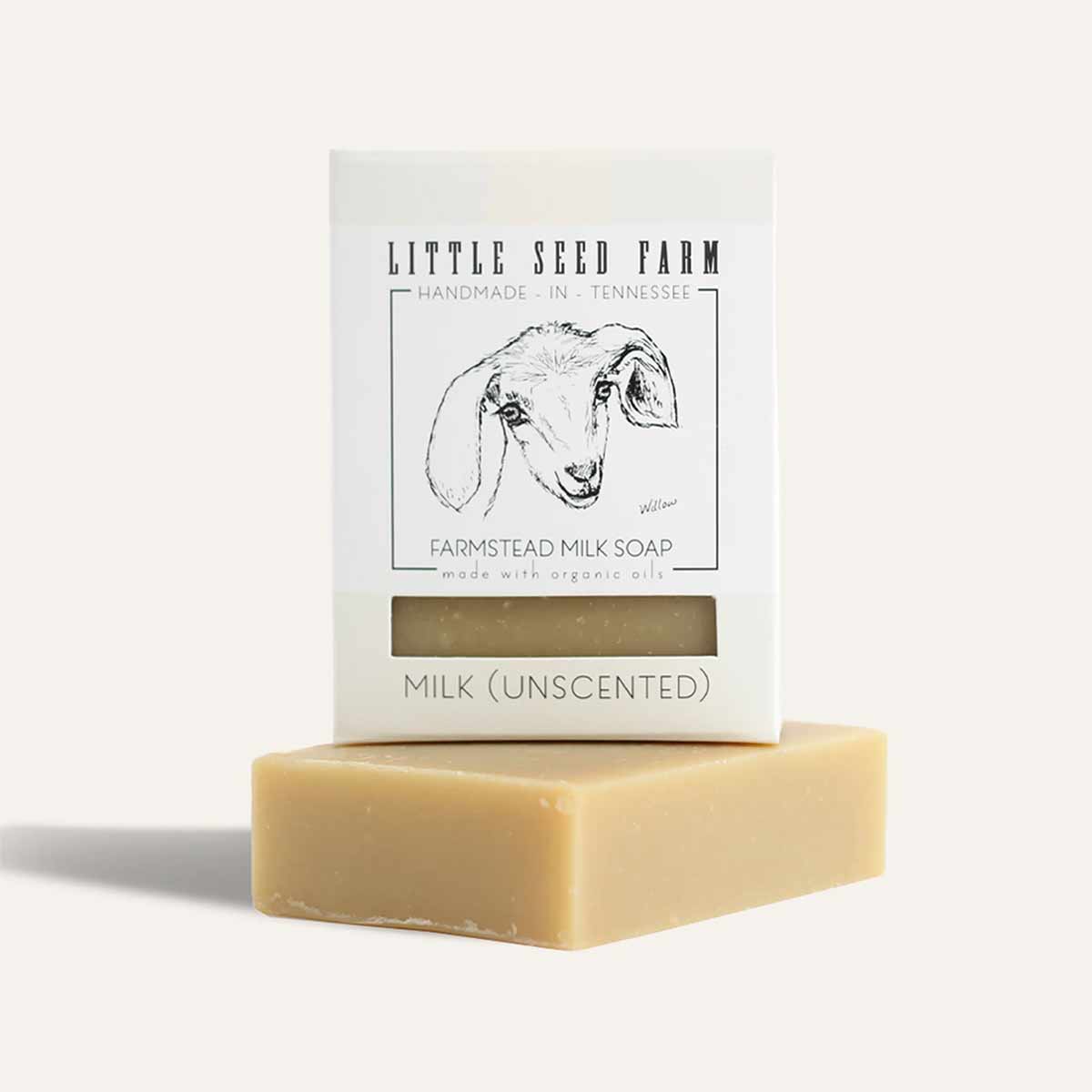 A cream-colored soap bar rests beneath a boxed bar with a goat illustration and a label reading Little Seed Farm milk soap.