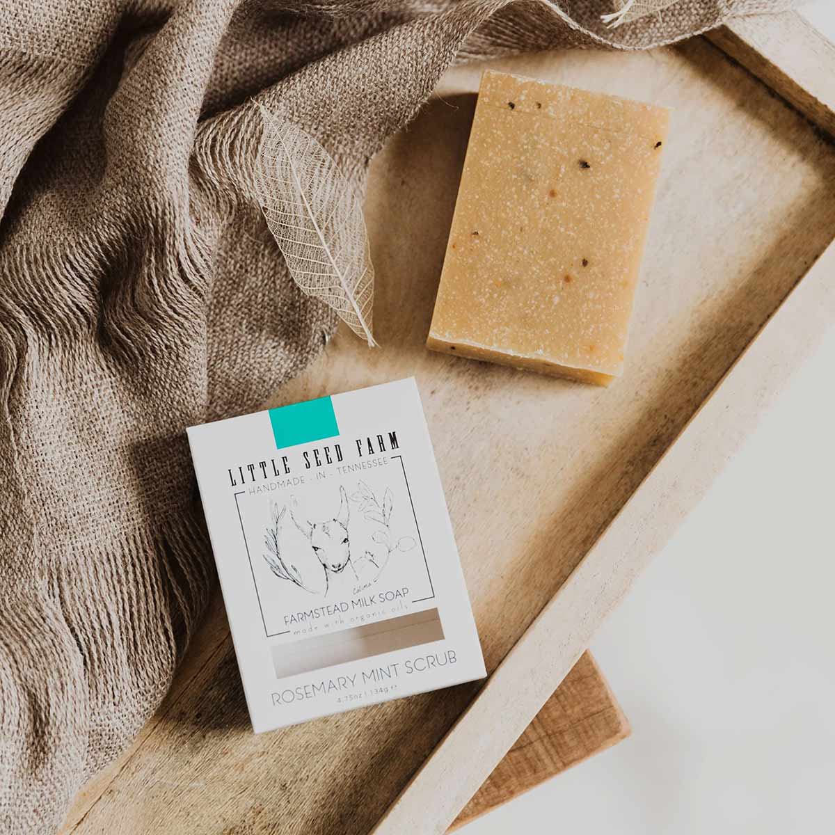 A speckled tan bar of soap rests beside its white Little Seed Farm Rosemary Mint Scrub box on a wooden tray with linen.