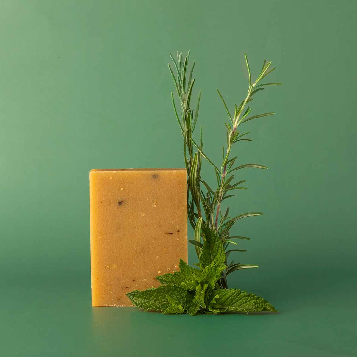 A rectangular golden-yellow bar of soap with small flecks stands upright beside fresh rosemary and mint sprigs against a green background.