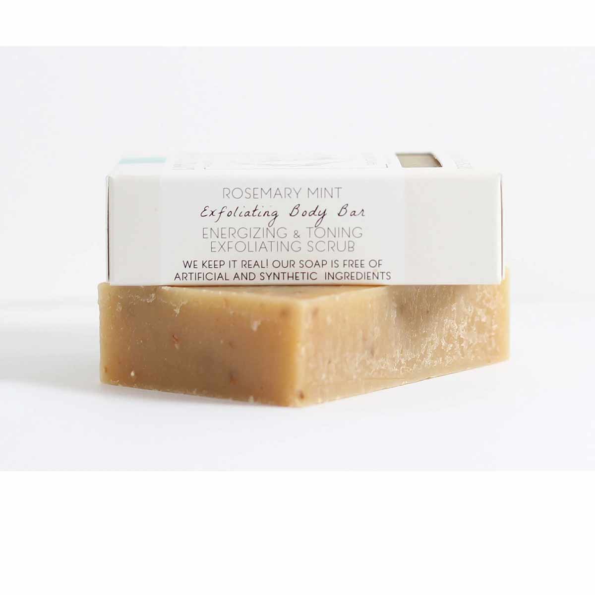 A tan speckled bar of soap sits beneath a white paper-wrapped bar labeled Rosemary Mint Exfoliating Body Bar.