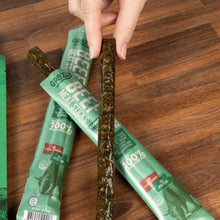 A hand holds an unwrapped herb-flecked beef meat stick above two green Grazly packages resting on a wooden table.