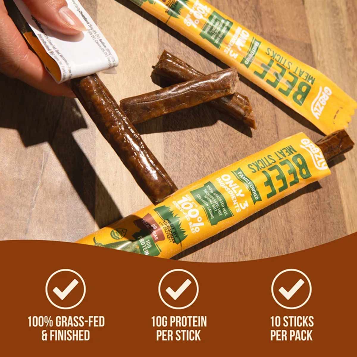 A hand peels back yellow Grazly beef meat stick wrappers on a wooden table, revealing several brown sticks nearby.