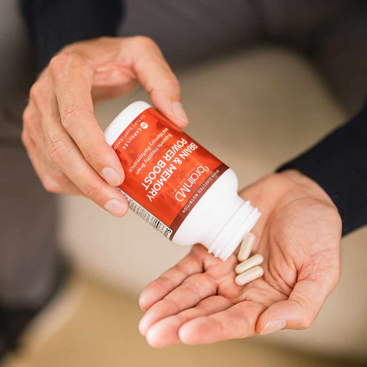Hand holding a red and white BrainMD supplement bottle with capsules pouring into palm.