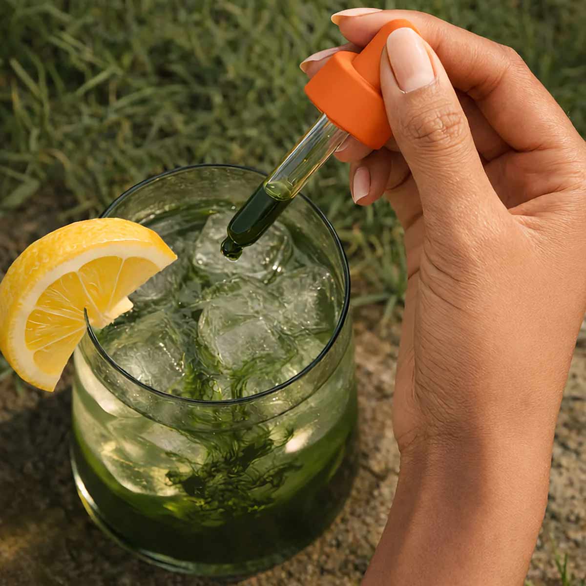 Dropper bottle with orange cap being held above a glass of green liquid with lemon slice and ice.