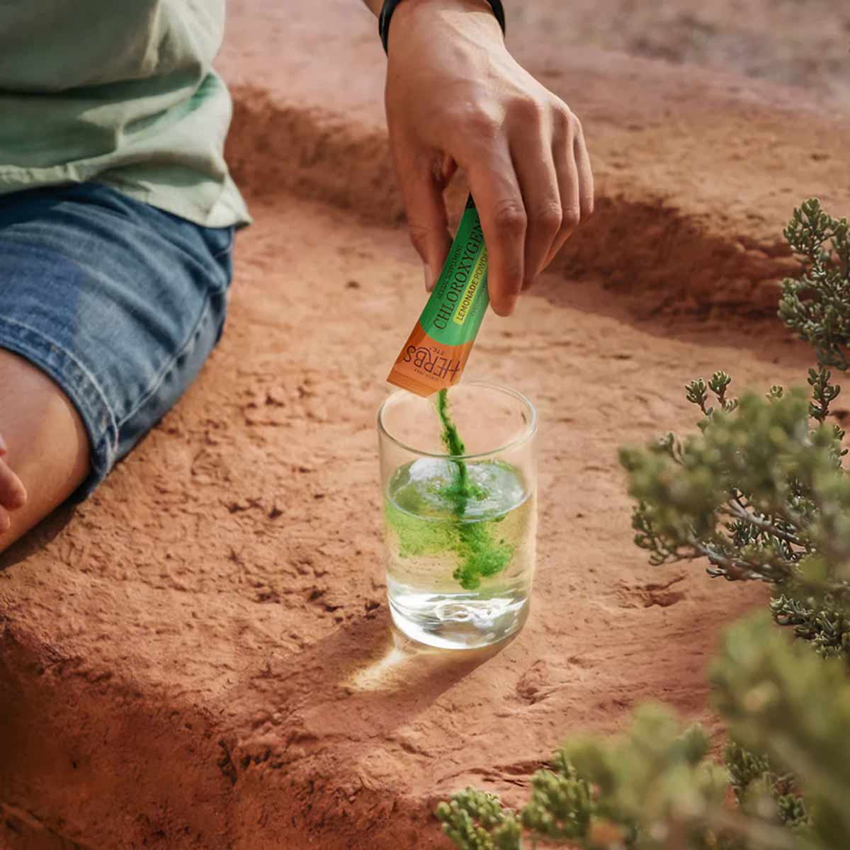 Person pouring green powder from packet into glass of water outdoors on sandy ground.