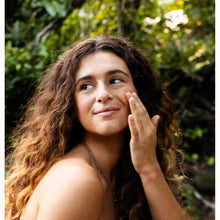 Woman with curly brown hair applying clear serum to her cheek against a leafy green background.