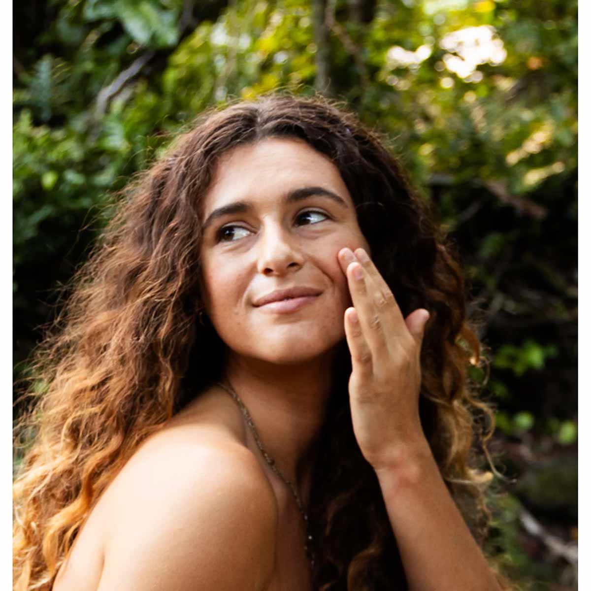 Woman with curly brown hair applying clear serum to her cheek against a leafy green background.