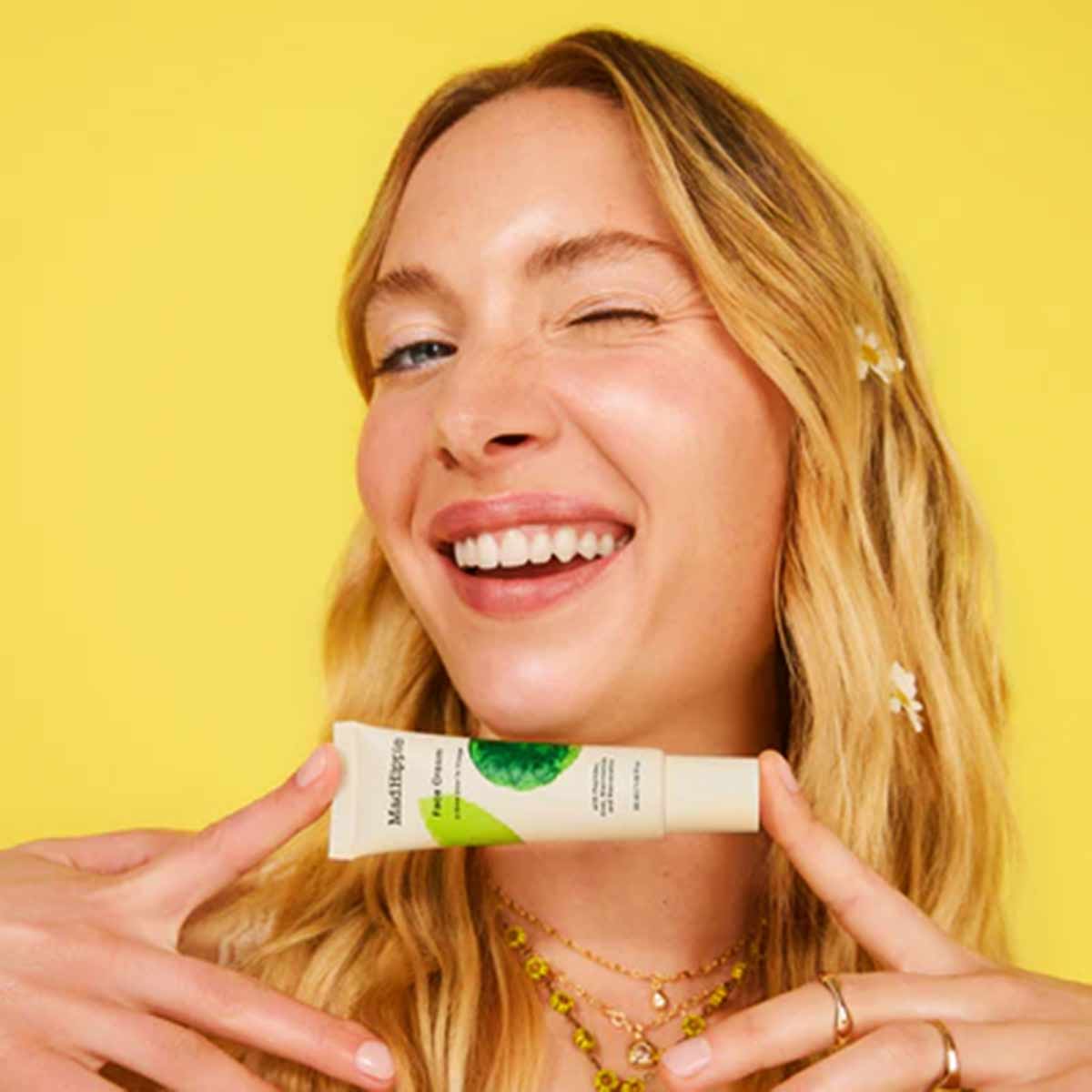 Woman with reddish-blonde hair holding a white Mad Hippie face cream tube with green accents against yellow background.
