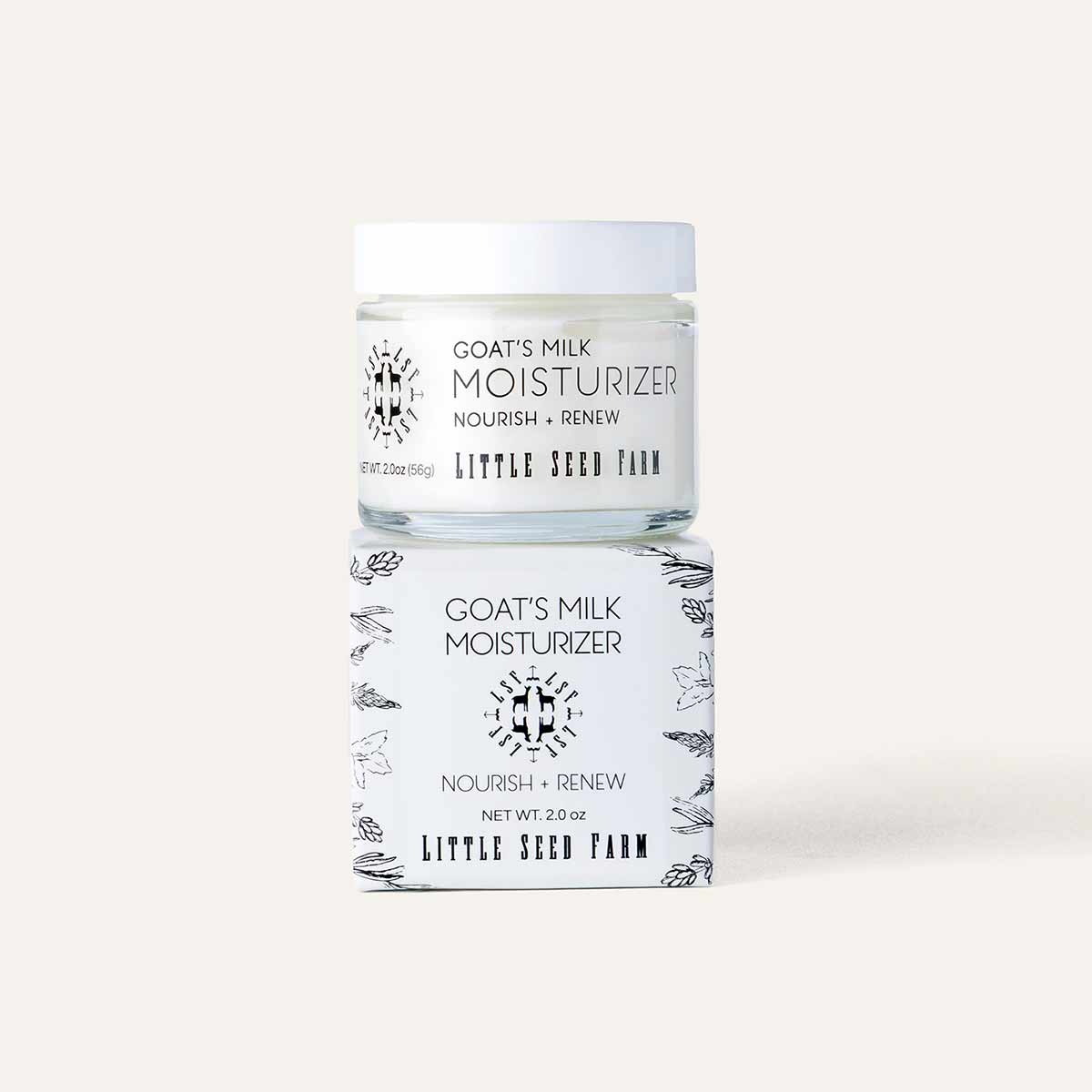 A glass jar of goat's milk moisturizer with a white lid rests on its white box decorated with botanical illustrations.