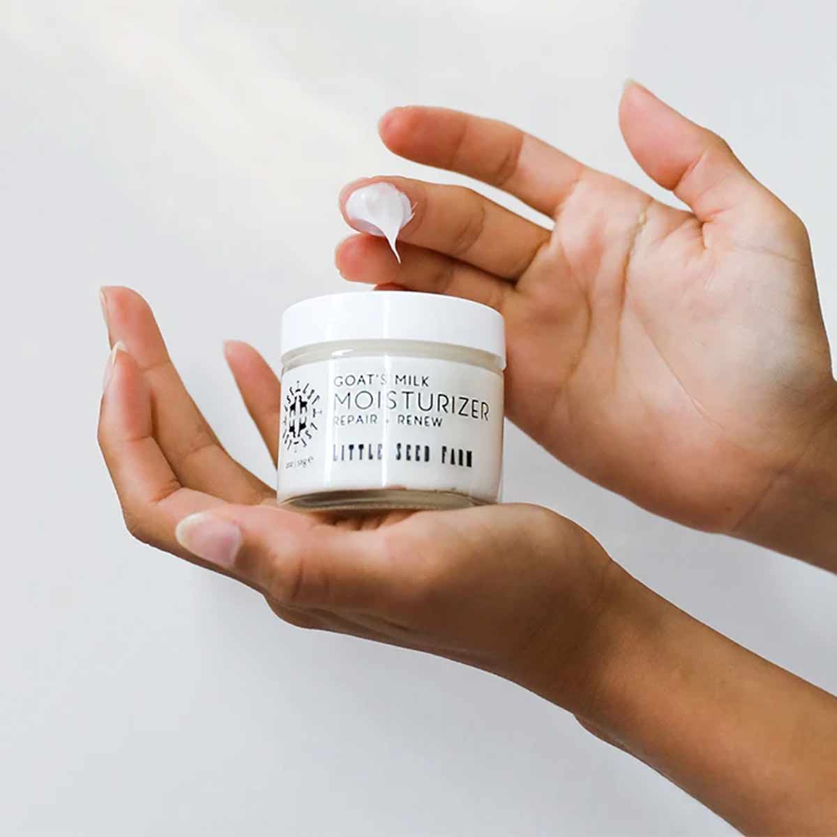 Two hands hold a small jar of Little Seed Farm goat's milk moisturizer while a fingertip lifts a dollop of white cream.