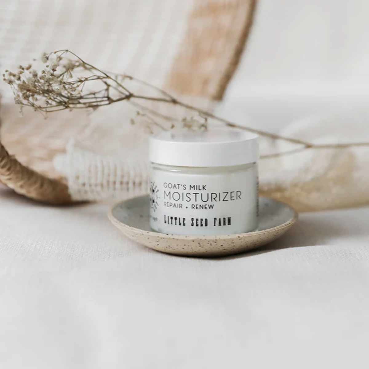 A small jar of Little Seed Farm goat's milk moisturizer with a white lid rests on a speckled ceramic dish beside dried flowers.