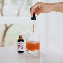 Hand holding dropper dispensing amber liquid from Lactivist bottle into glass of brown liquid.