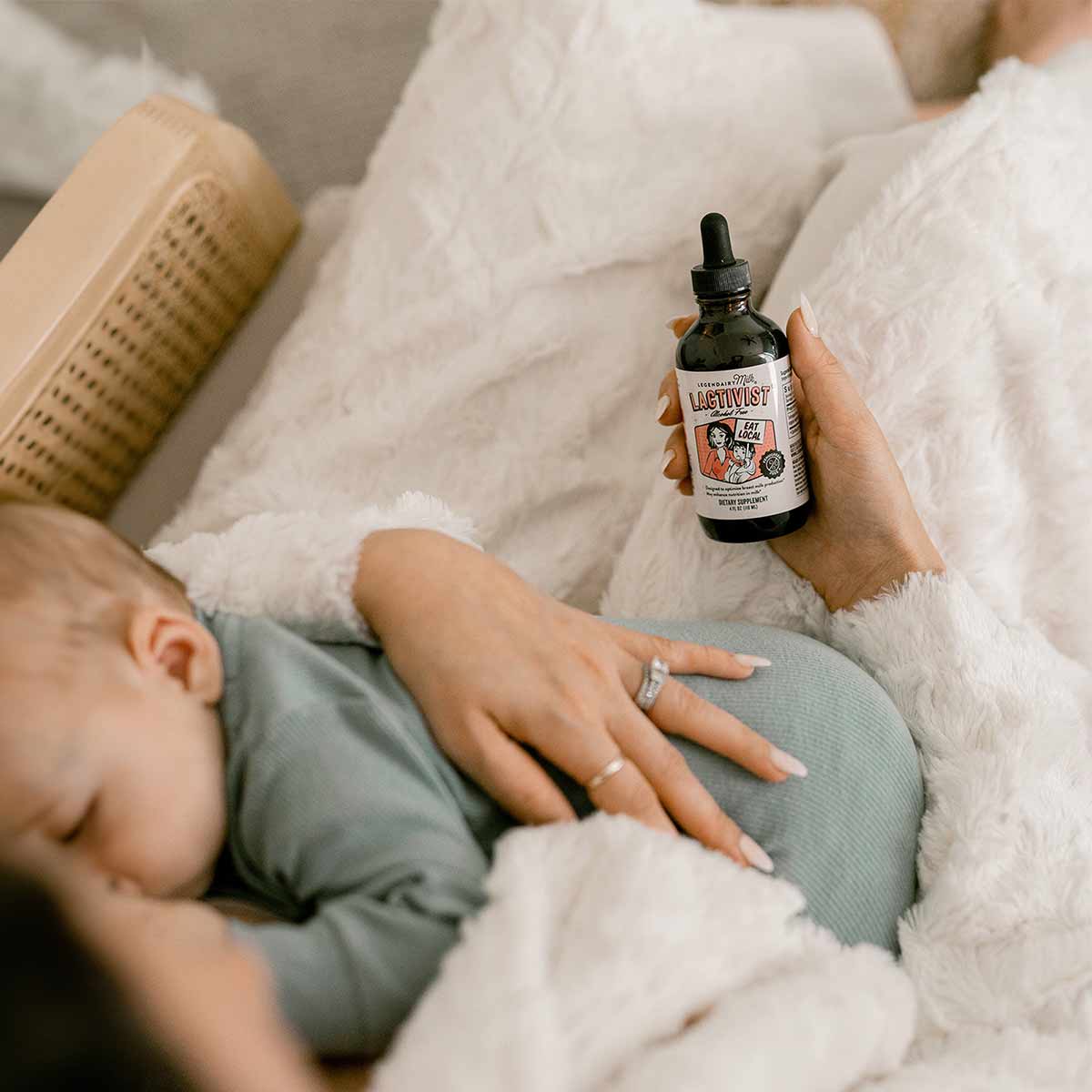 Person holding Lactivist dropper bottle with nursing baby and massage tool on white bedding.