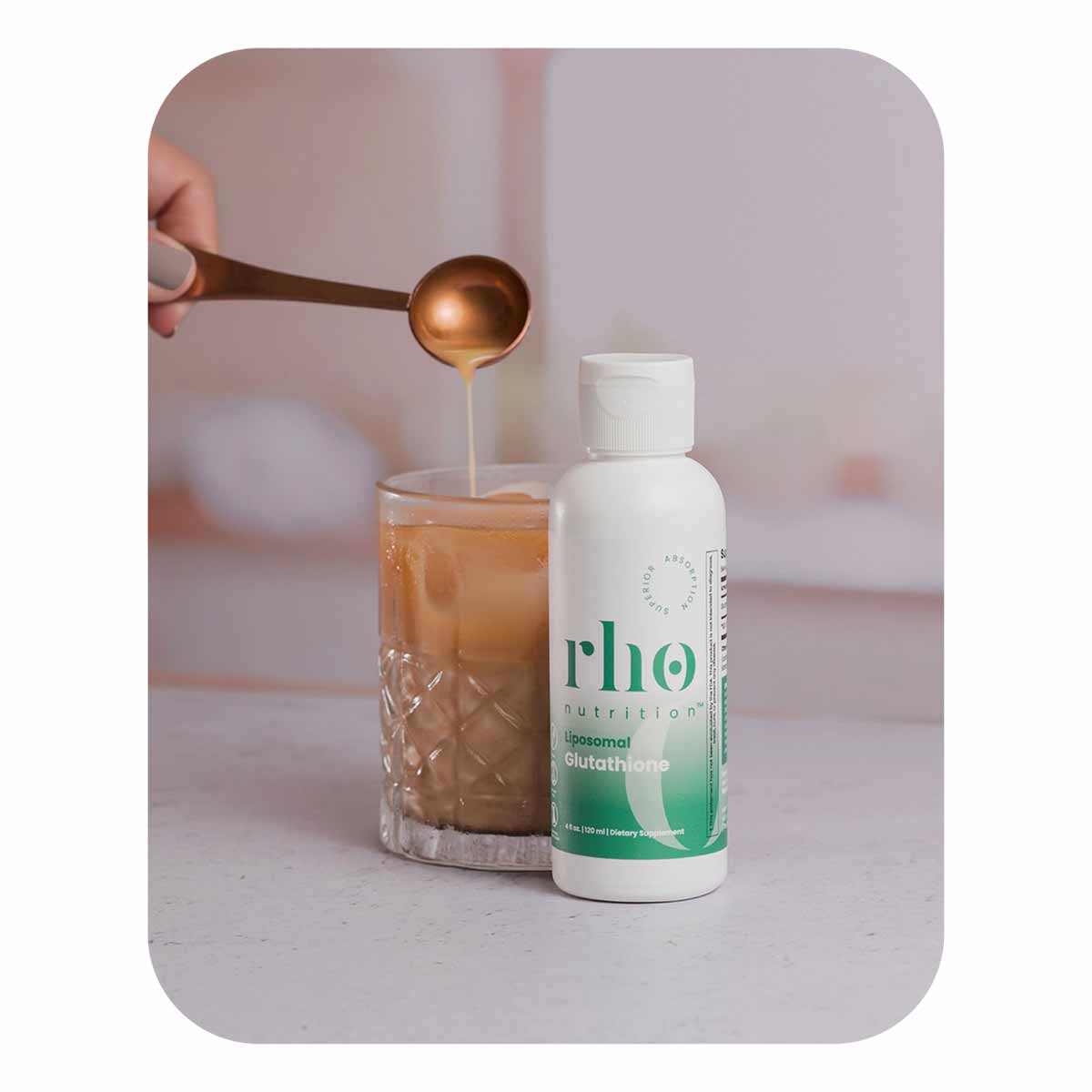 Rho Nutrition liposomal glutathione bottle next to glass with golden liquid being drizzled from bronze measuring spoon.