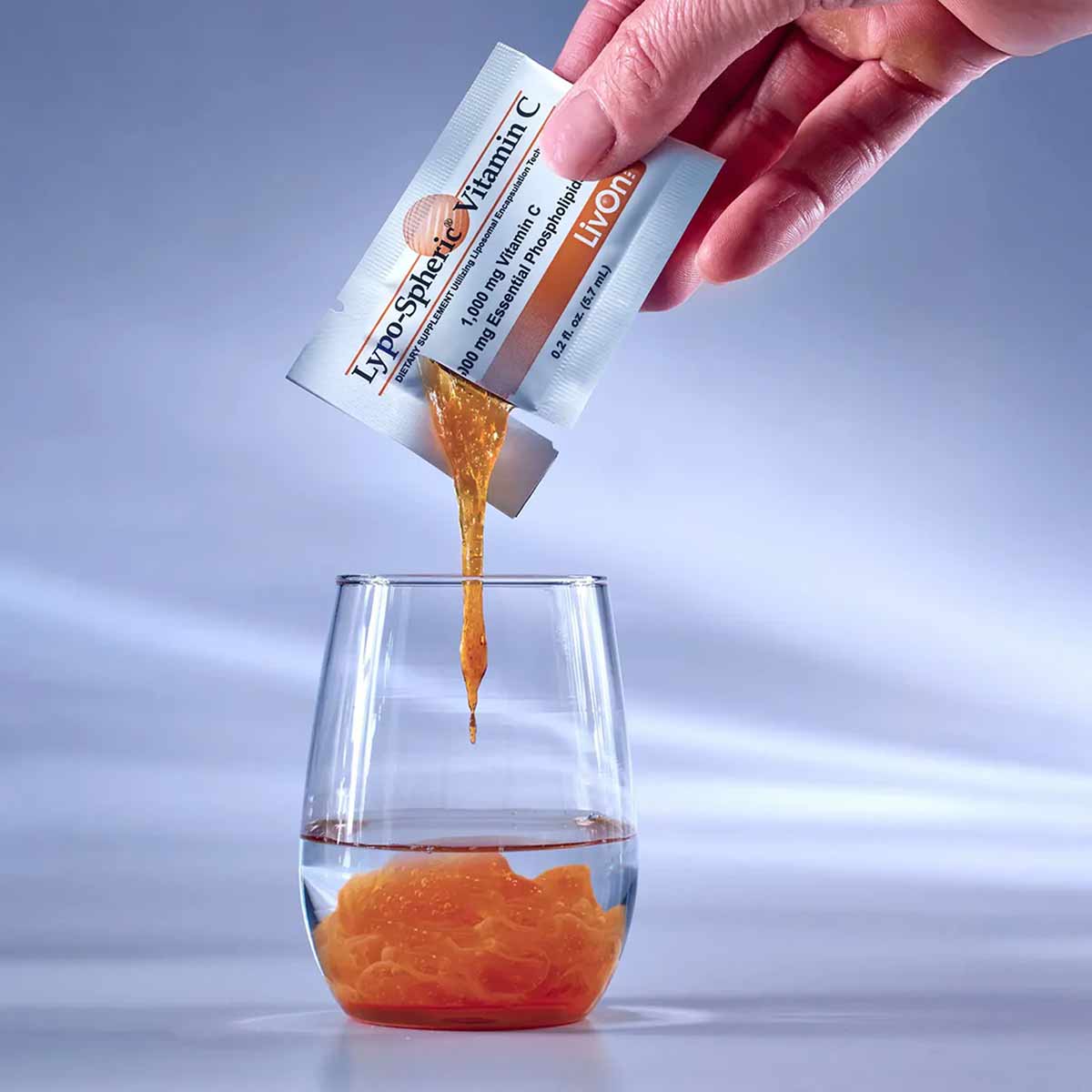 Hand squeezing Lypo-Spheric Vitamin C packet, dispensing golden liquid into glass.