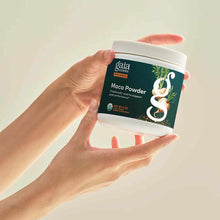 Hand holding Gaia Herbs Maca Powder jar with dark teal label and white screw-top lid against neutral background.