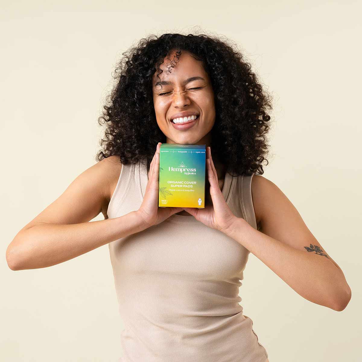 A smiling woman with curly dark hair holds a green and yellow box of Hempress Organic Cover Super Pads against a cream background.