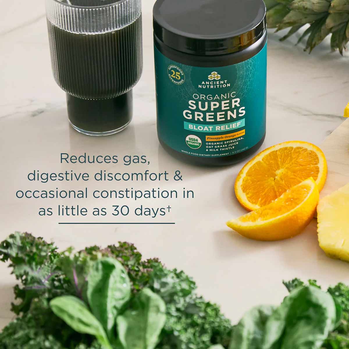 Teal container of Organic SuperGreens + Bloat Relief powder with glass of green beverage, orange slices, and fresh kale.