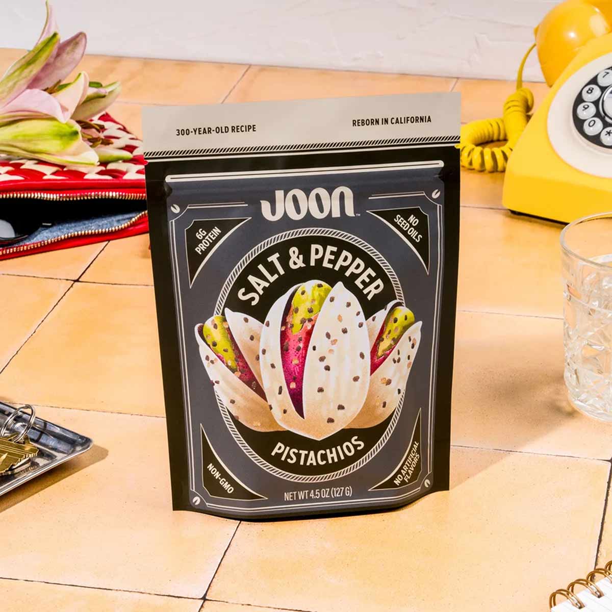 Black Joon brand pouch of salt and pepper pistachios on a wooden surface surrounded by vintage decor.