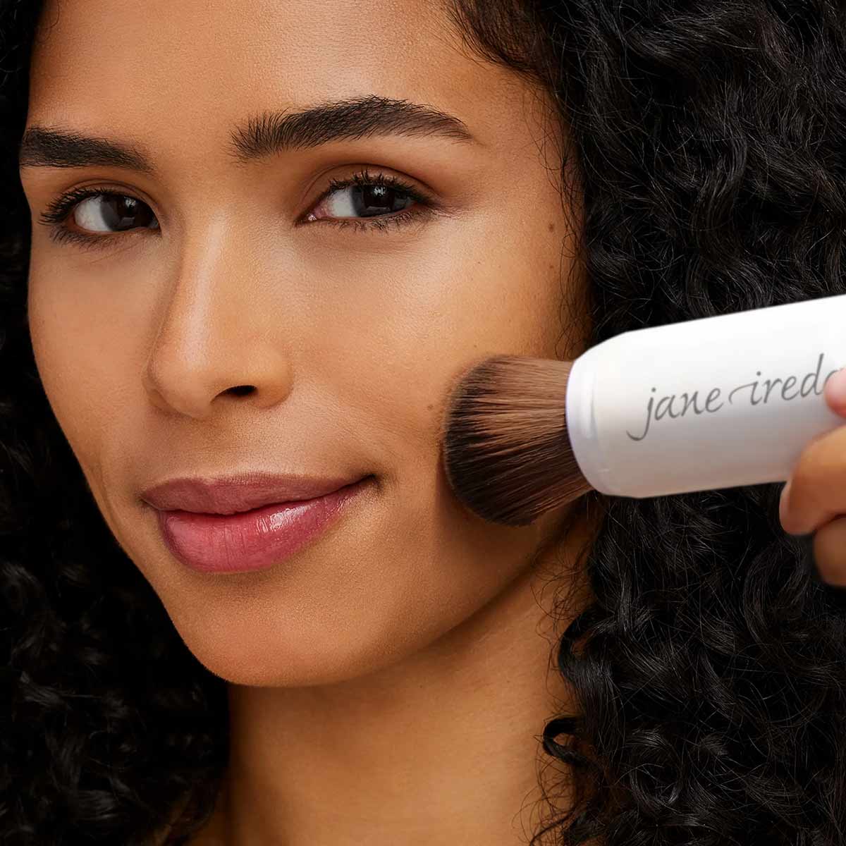 White cylindrical brush applicator with jane iredale branding being applied to a woman's cheek.