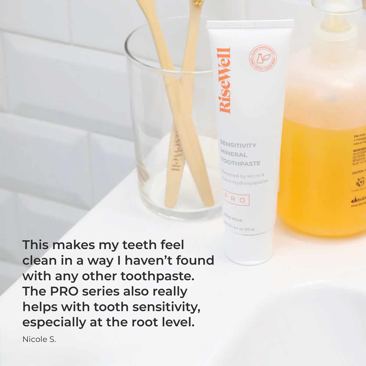 RiseWell mineral toothpaste tube next to bamboo toothbrushes in glass holder on white bathroom counter.
