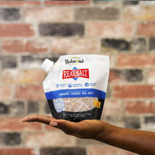 White pouch of Real Salt coarse sea salt by Redmond Life held in a hand against a brick wall background.