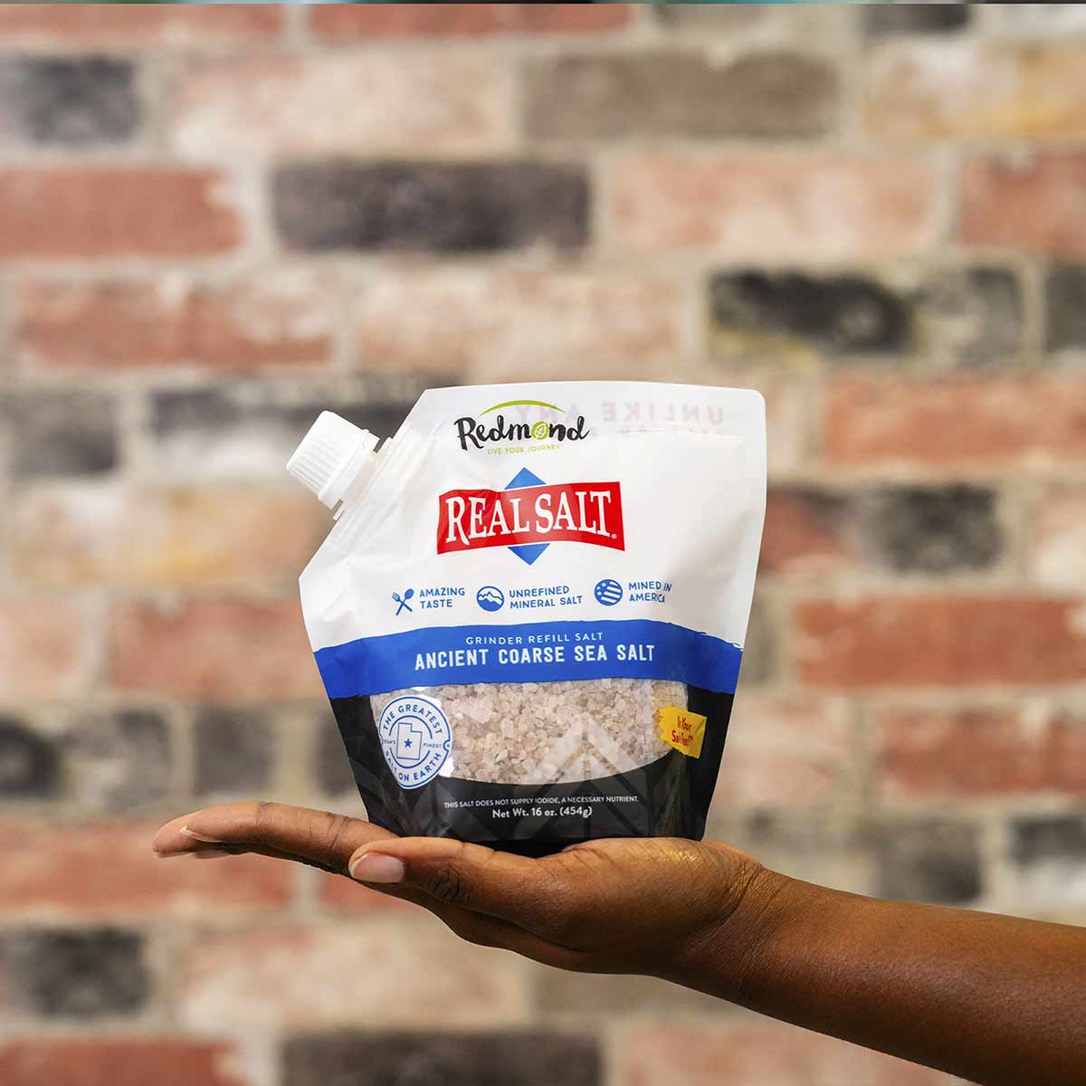 White pouch of Real Salt coarse sea salt by Redmond Life held in a hand against a brick wall background.