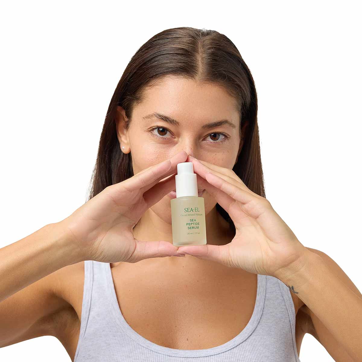 A woman in a gray tank top holds a small frosted bottle of Sea-El Sea Peptide Serum between her hands.
