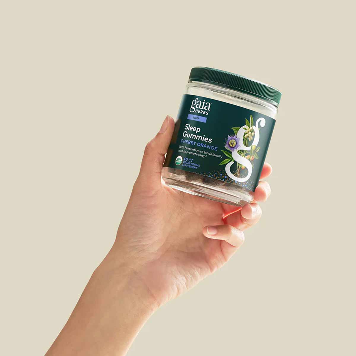 Hand holding a clear glass jar of Gaia Herbs Sleep Gummies in Cherry Orange flavor with a dark green lid.