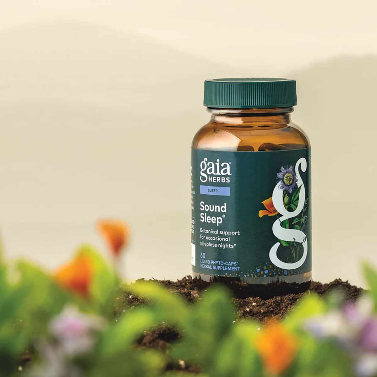 Gaia Herbs Sound Sleep bottle with teal cap and label, sitting on soil with blurred flowers in background.