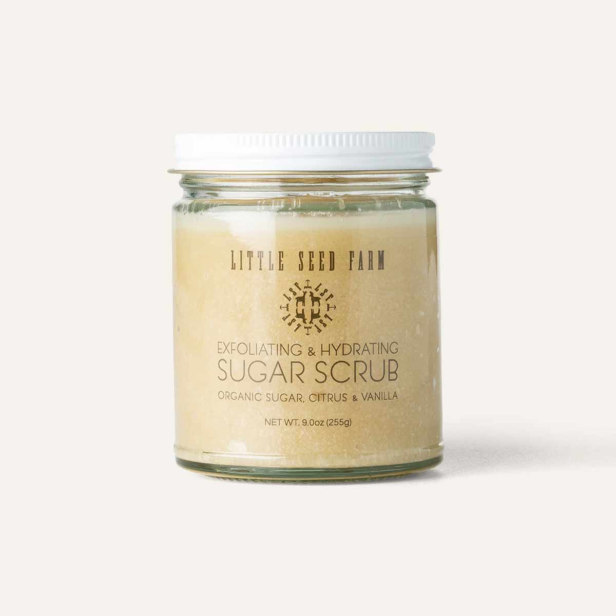 A clear glass jar with a white lid holds pale golden sugar scrub, labeled Little Seed Farm Exfoliating & Hydrating Sugar Scrub.